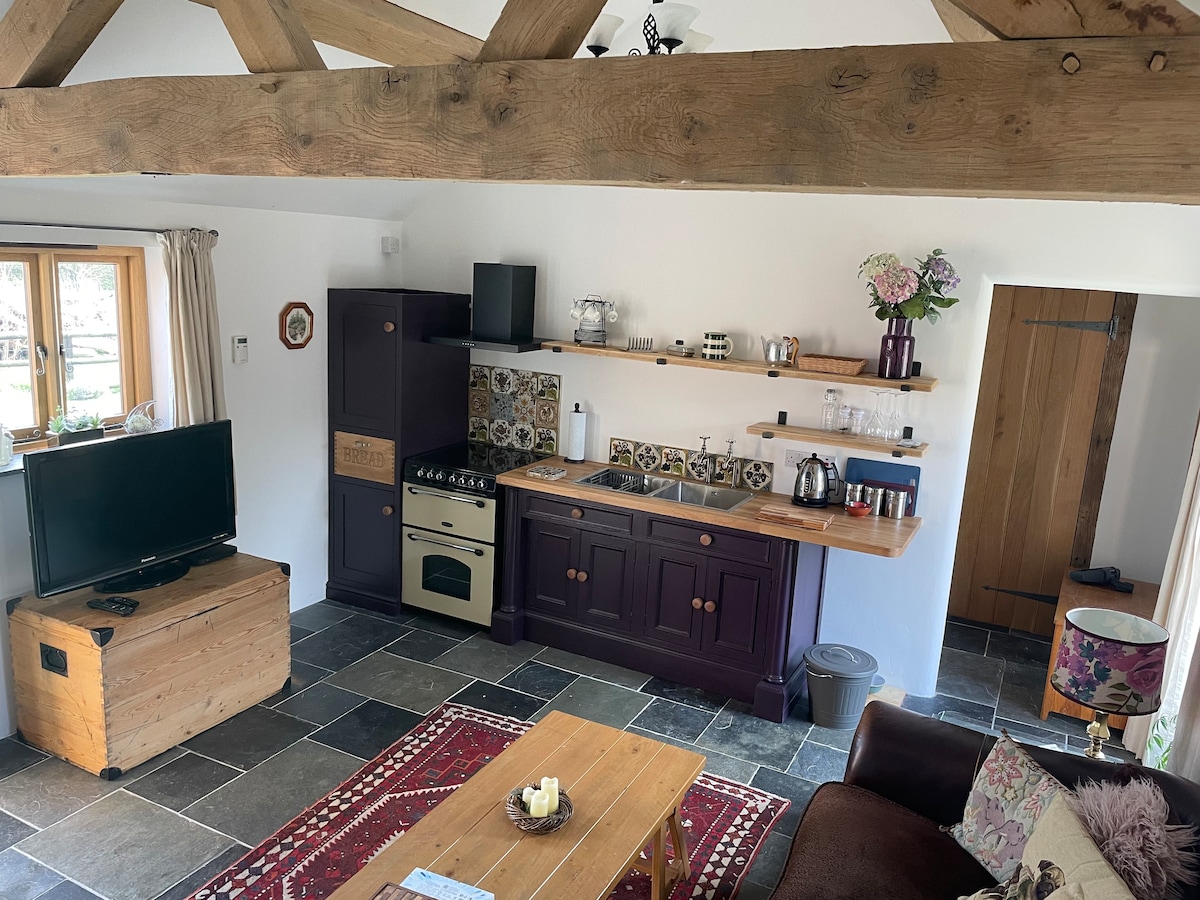 The open-plan kitchen and living area features a wooden ceiling with exposed beams. A cozy seating arrangement includes a sofa, accompanied by a coffee table. The kitchen is equipped with a stove, refrigerator, and wooden shelving displaying kitchenware. Natural light from windows brightens the space.