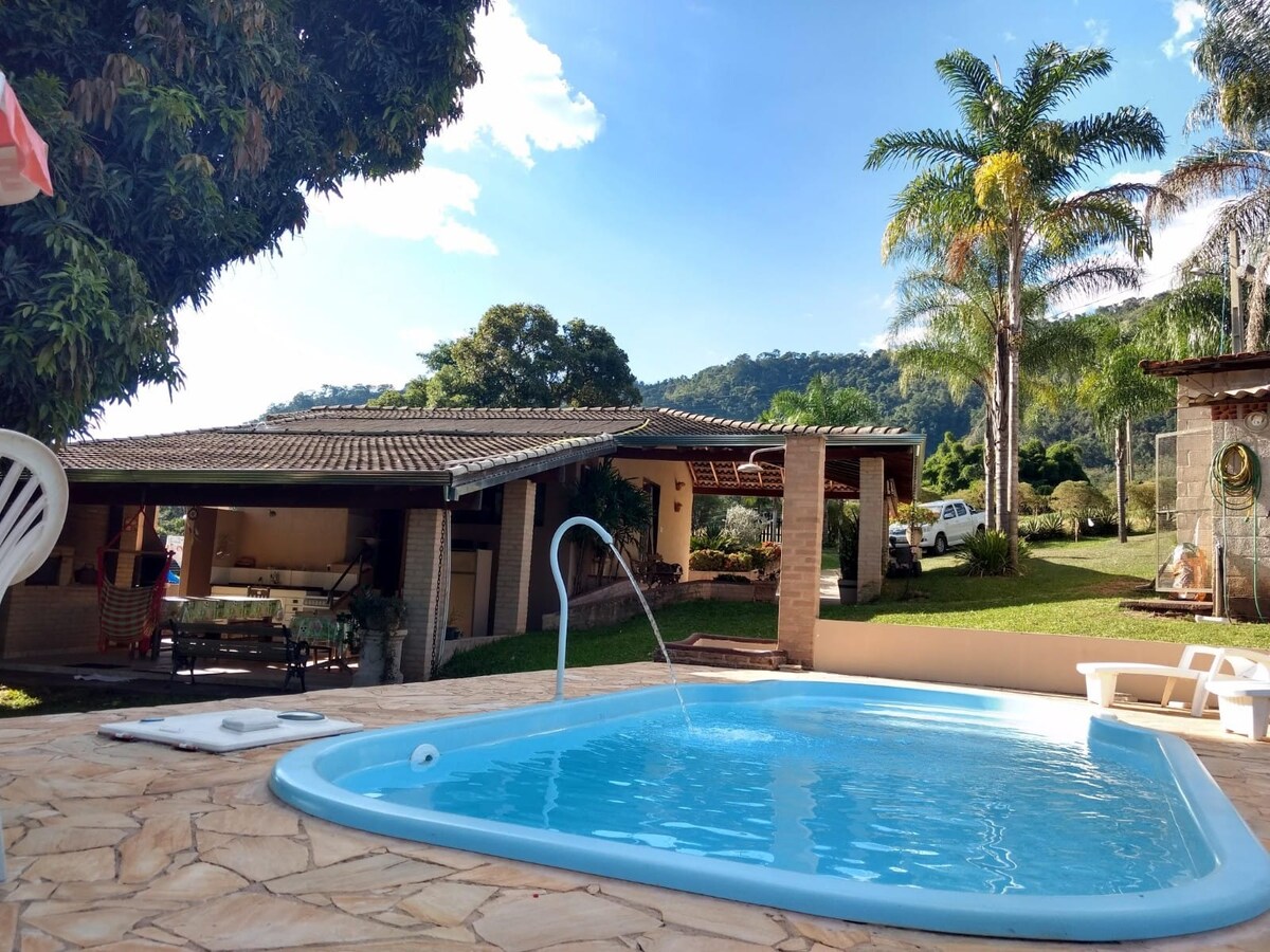 A clear blue pool is visible in the foreground, surrounded by a patio with natural stone flooring. In the background, a serene landscape of green hills and trees contrasts with a single-story residence featuring a shaded outdoor area and ample open space.
