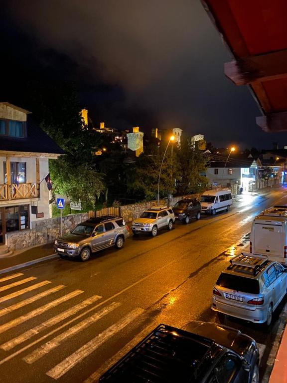 A nighttime view of a bustling street scene, showcasing parked vehicles lined along a wet road. Streetlights illuminate the surroundings, and trees are visible alongside the buildings. In the distance, illuminated structures can be seen on a hillside, adding depth to the cityscape.