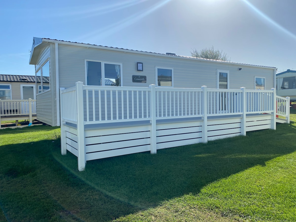A modern caravan is presented with a bright exterior and a white railing surrounding the spacious decking area. Lush green grass frames the base, while large windows allow for ample natural light. The structure is well-maintained, showcasing a welcoming outdoor space.