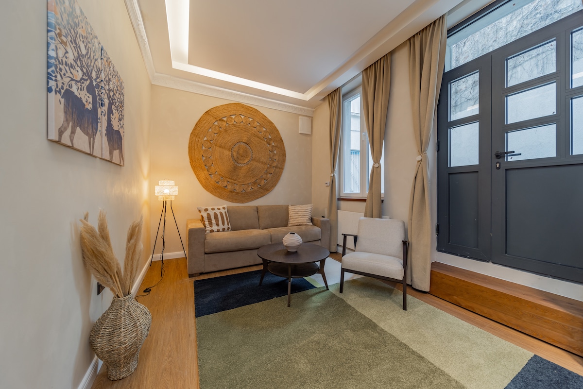 A cozy living area features a comfortable sofa and a round coffee table positioned on a cozy area rug. Soft lighting is provided by a floor lamp beside the couch, and natural light enters through large doors adorned with decorative curtains.