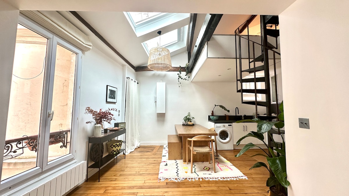 The bright living area features a large wooden table surrounded by four chairs, with a fully equipped kitchen visible. A spiral staircase leads to the mezzanine sleeping area. Skylights above allow natural light to fill the space, while potted plants add a touch of greenery.