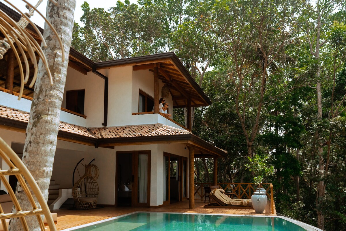 The villa is nestled among trees, with a blend of natural materials. A large wooden deck surrounds a private swimming pool, inviting relaxation. Large windows provide views of the lush surroundings, enhancing the connection with nature. A hanging chair is positioned for comfortable seating.