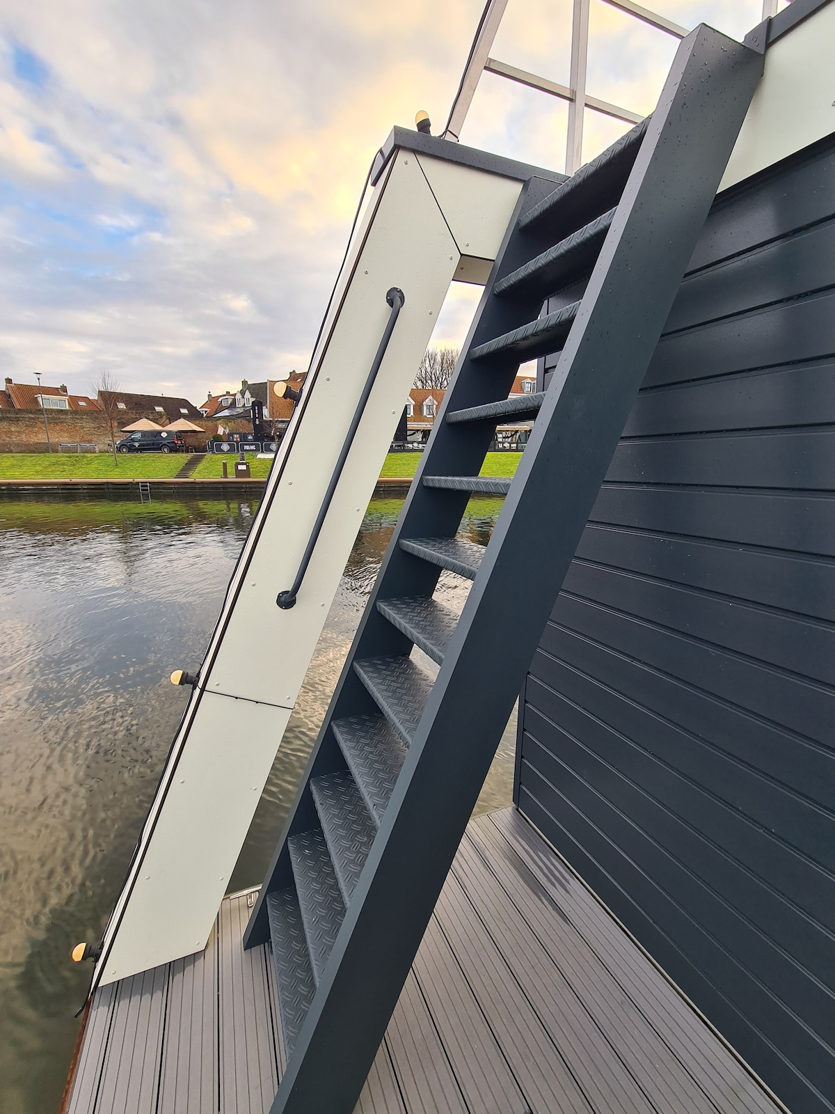 A sturdy staircase is visible, leading from the deck of the houseboat. The steps are securely attached, and the railing offers support. The setting includes tranquil water alongside the shore, with buildings visible in the background.