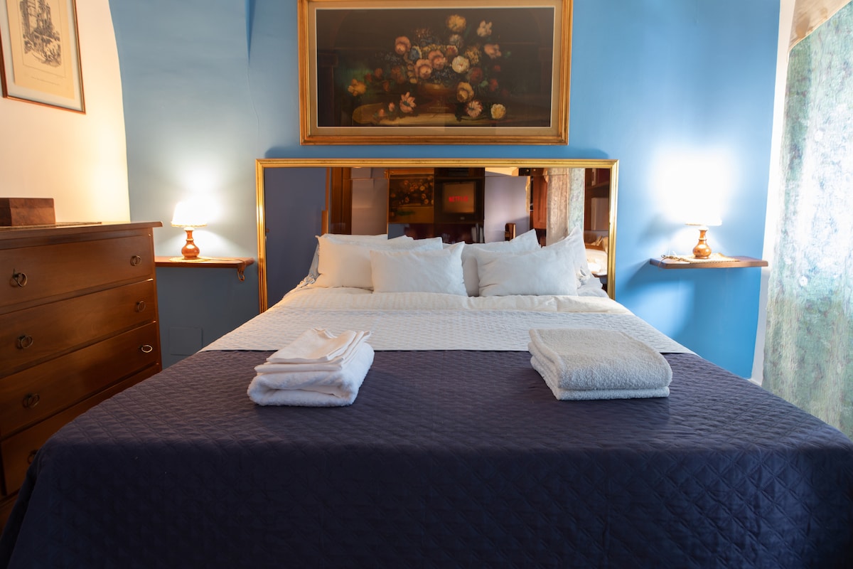 A spacious bed with a navy blue coverlet and white linens is situated against a blue wall. Decorative white pillows are arranged at the head of the bed. The room is softly illuminated by lamps on each side, with a piece of artwork above the bed. A dresser is visible to the left.