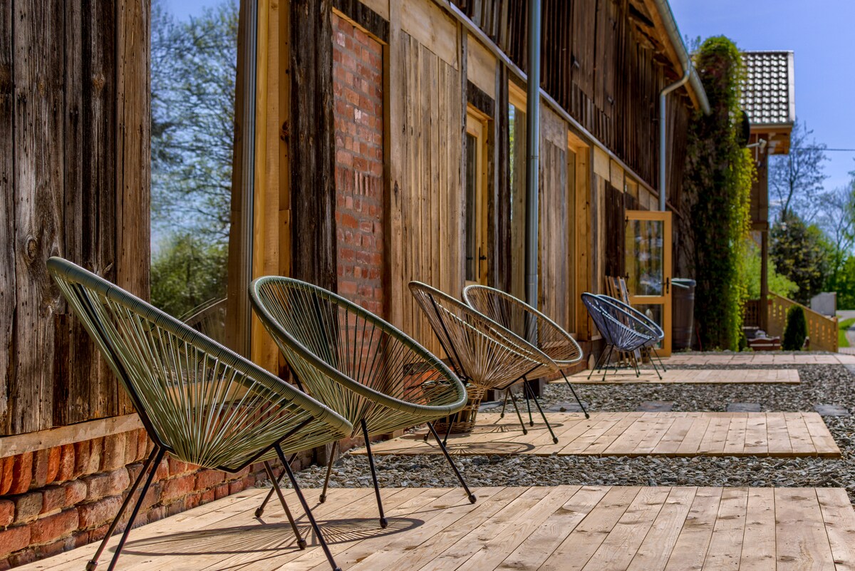 A row of four woven chairs is positioned on a wooden deck, offering a space to relax outdoors. The rustic wooden structure serves as a backdrop, featuring large glass doors that enhance the connection to the surrounding nature.