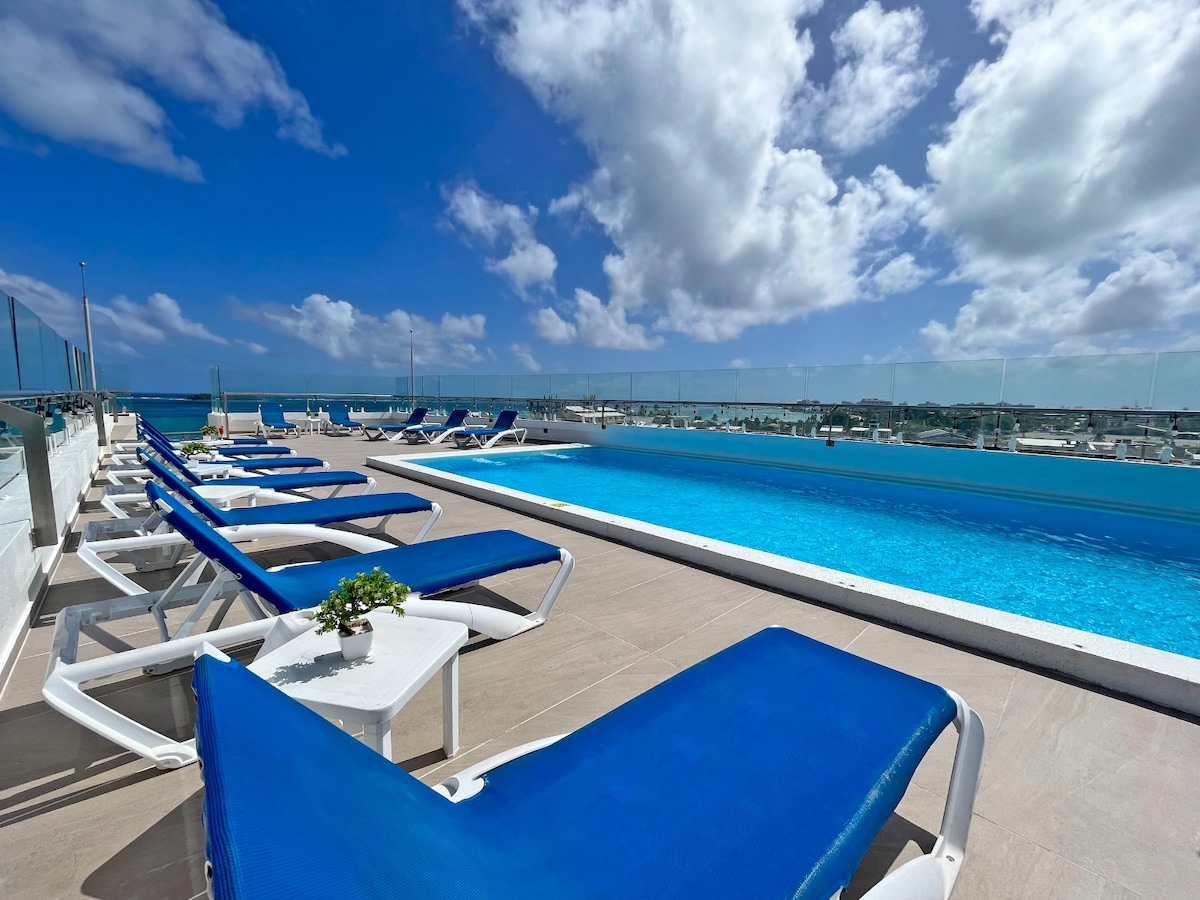 A rooftop pool area is shown, featuring lounge chairs arranged around an inviting blue pool. Clear views of the sky and distant shoreline are visible, with white clouds scattered above. A small potted plant sits on a side table beside the lounge chairs.