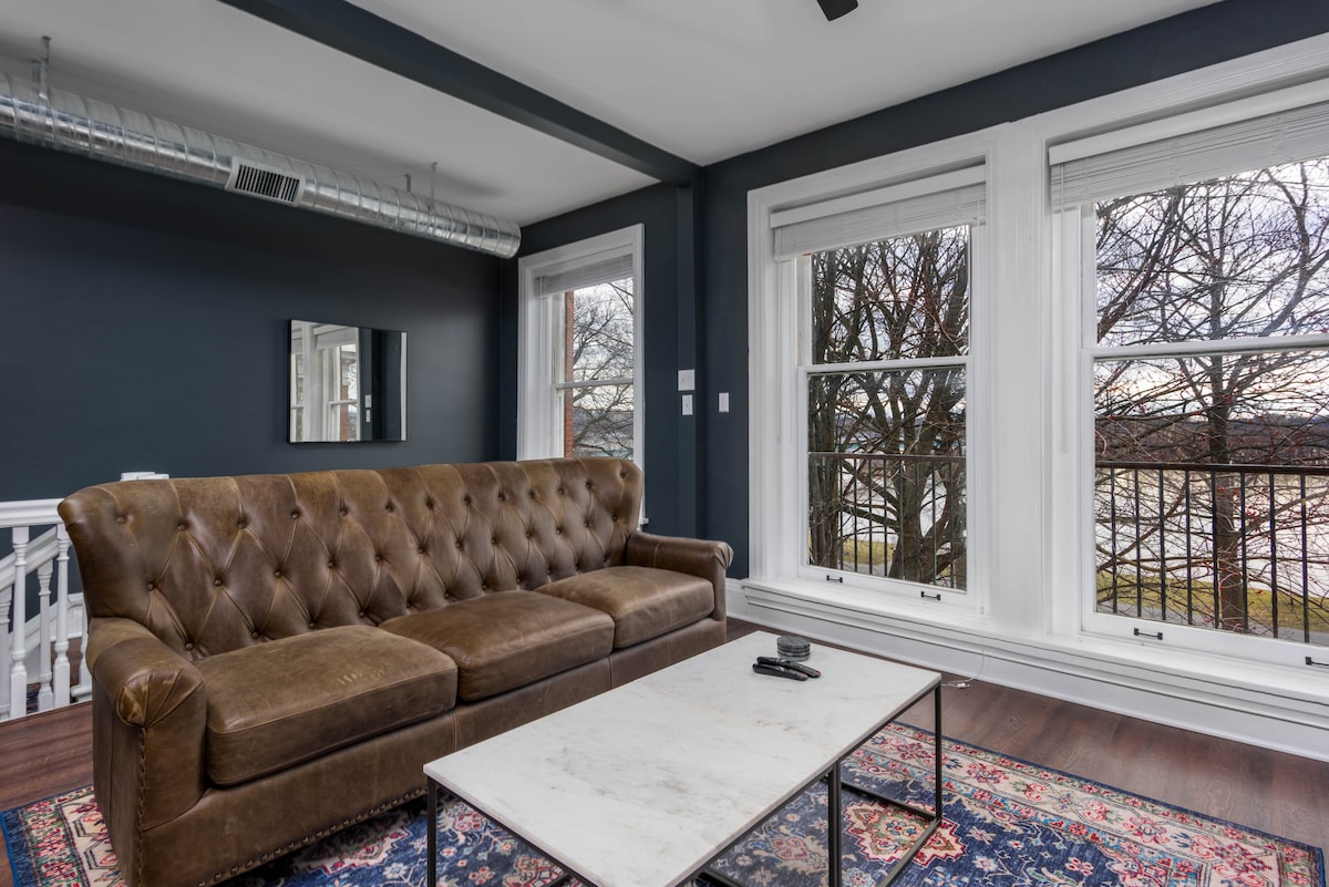A spacious living area is featured with a brown tufted leather sofa positioned across from a sleek marble coffee table. Large windows provide views of the surrounding trees, while dark accent walls create a modern aesthetic.