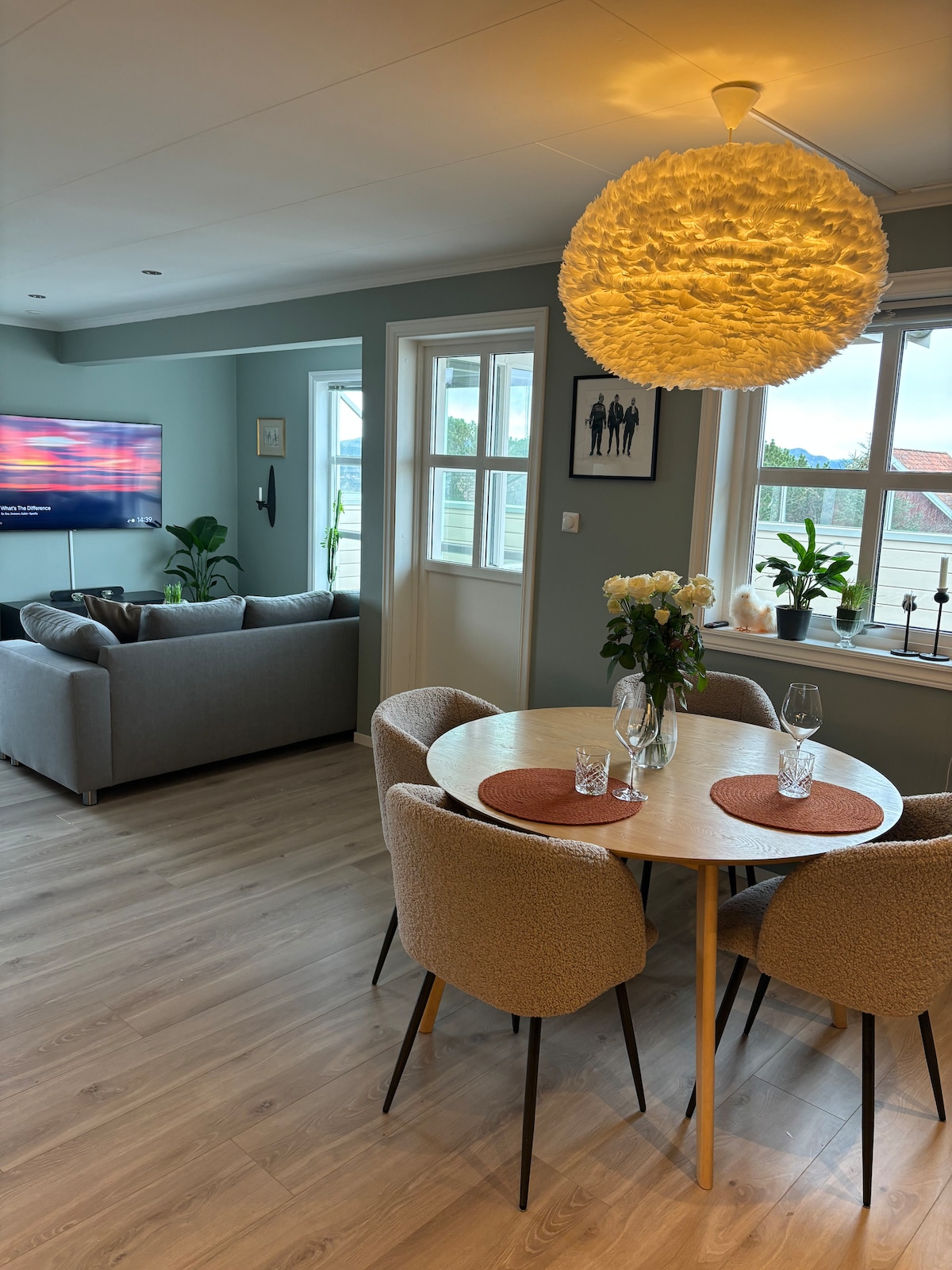 A dining area features a round table surrounded by four upholstered chairs. A bouquet of flowers sits at the center, complemented by glassware. In the background, a television is mounted on the wall, with comfortable seating visible nearby. Large windows allow natural light to fill the space.