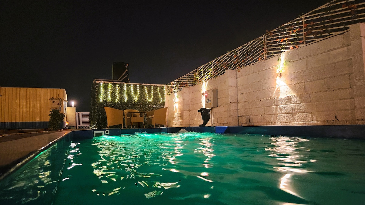 An illuminated private swimming pool glimmers under the night sky, surrounded by a stone wall adorned with greenery. Soft light fixtures enhance the serene atmosphere. Poolside, a small seating area with chairs and a table invites relaxation.