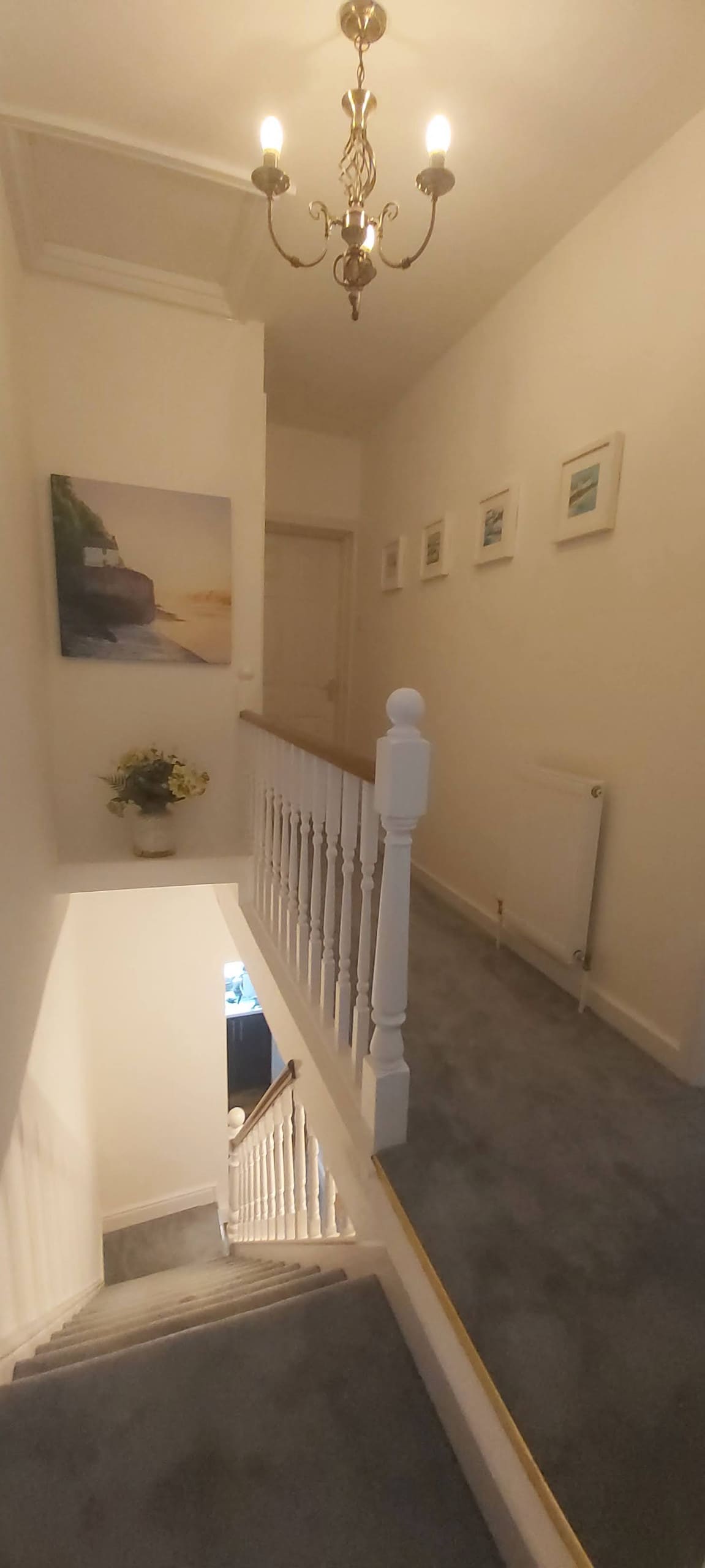 A well-lit hallway features neutral walls and carpet, leading to an upper landing. A small piece of wall art depicting a coastal scene hangs above a decorative plant. Framed pictures line the walls, contributing to a clean and inviting passage.