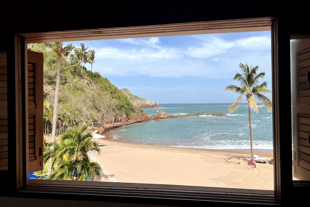A scenic view through open wooden shutters reveals a sandy beach bordered by lush palm trees and rocky formations. Soft waves gently lap at the shore under a partly cloudy sky, creating a serene coastal atmosphere.