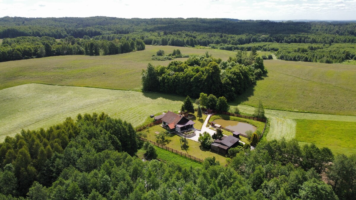 Aerial view of a serene property surrounded by lush green fields and dense forests. Various buildings and a winding pathway are visible within the expansive landscape, which offers a sense of openness and tranquility.