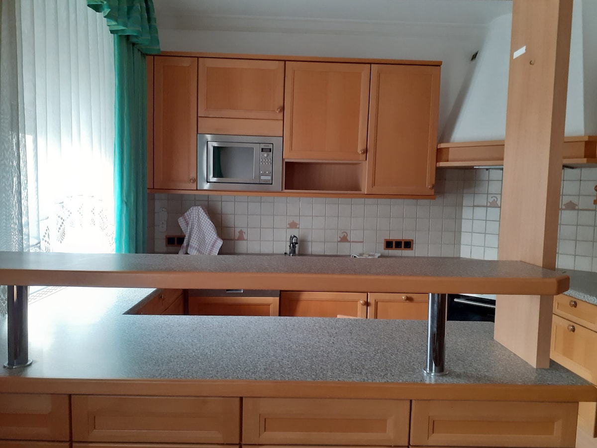 A functional kitchen features wooden cabinetry and a granite countertop. Essential appliances, including a microwave, are integrated into the design. A window with sheer curtains allows natural light to filter in, enhancing the bright and open feel of the space.