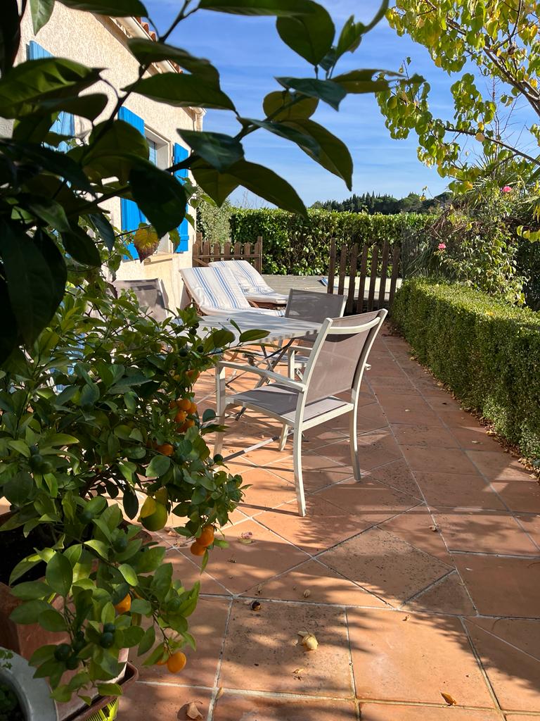 A sunlit patio area features several light-colored lounge chairs and a small table, surrounded by vibrant greenery and flowering plants. The terracotta tiles create a warm ambiance, while a wooden fence provides a sense of privacy and separation from surrounding spaces.