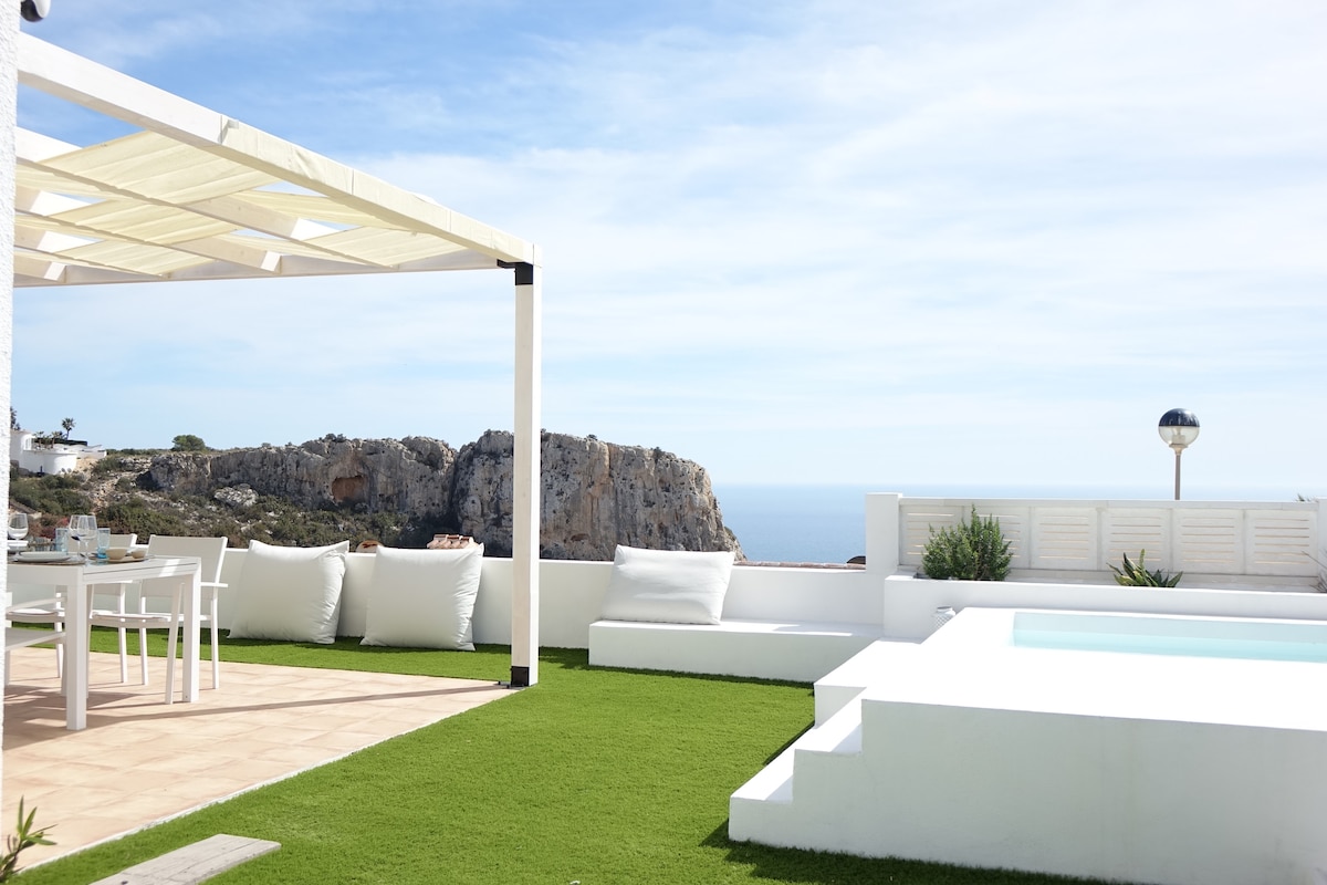 An outdoor terrace is featured, with a dining area under a shaded pergola. Comfortable outdoor seating and a small pool are visible, set against a backdrop of the sea and distant cliffs. Lush green grass complements the overall serene space.