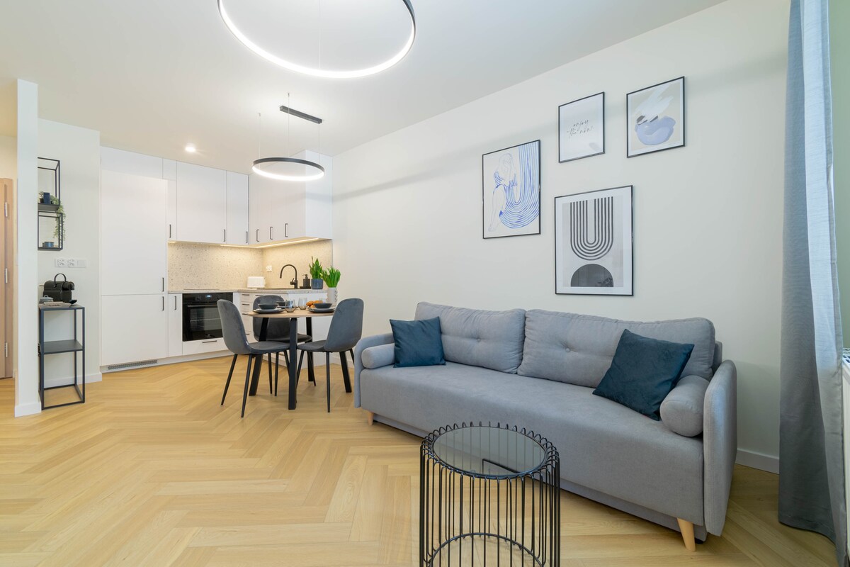 A modern living space features a light grey sofa complemented by dark cushions. A round black coffee table sits in front. Nearby, a dining area includes a round table with four chairs. Neutral wall colors are accented by framed art above the sofa.