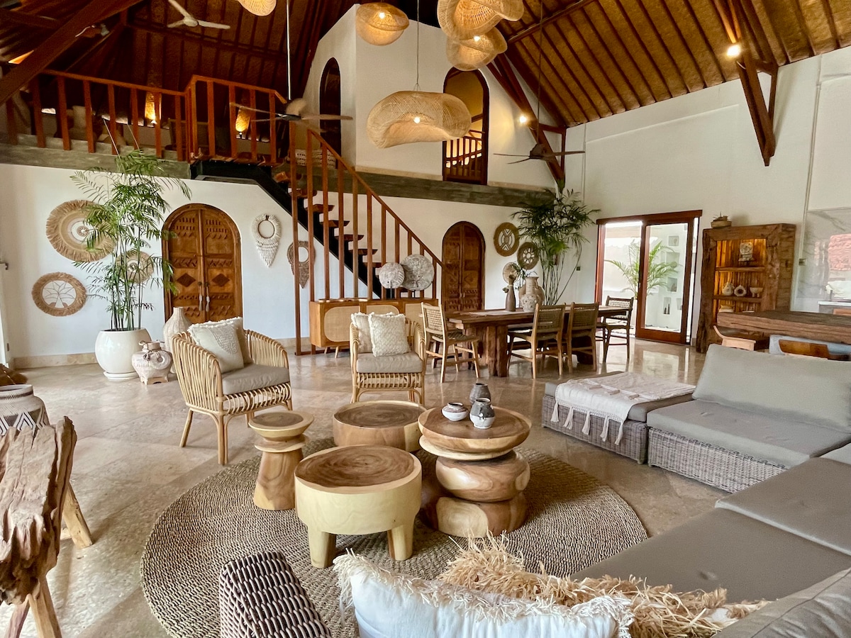 The spacious living area features high, wooden ceilings with natural light flooding through large windows. Various seating arrangements include woven chairs and a sectional sofa surrounding circular wooden tables. Decor elements highlight traditional Indonesian craftsmanship, complemented by lush greenery in the background.