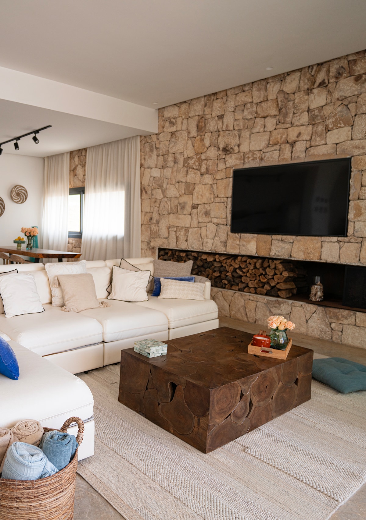 A light-filled living area features a white sectional sofa adorned with various cushions. A wooden coffee table sits centrally, complemented by decorative items. A textured stone wall serves as a backdrop, while firewood is neatly arranged beneath the entertainment system.