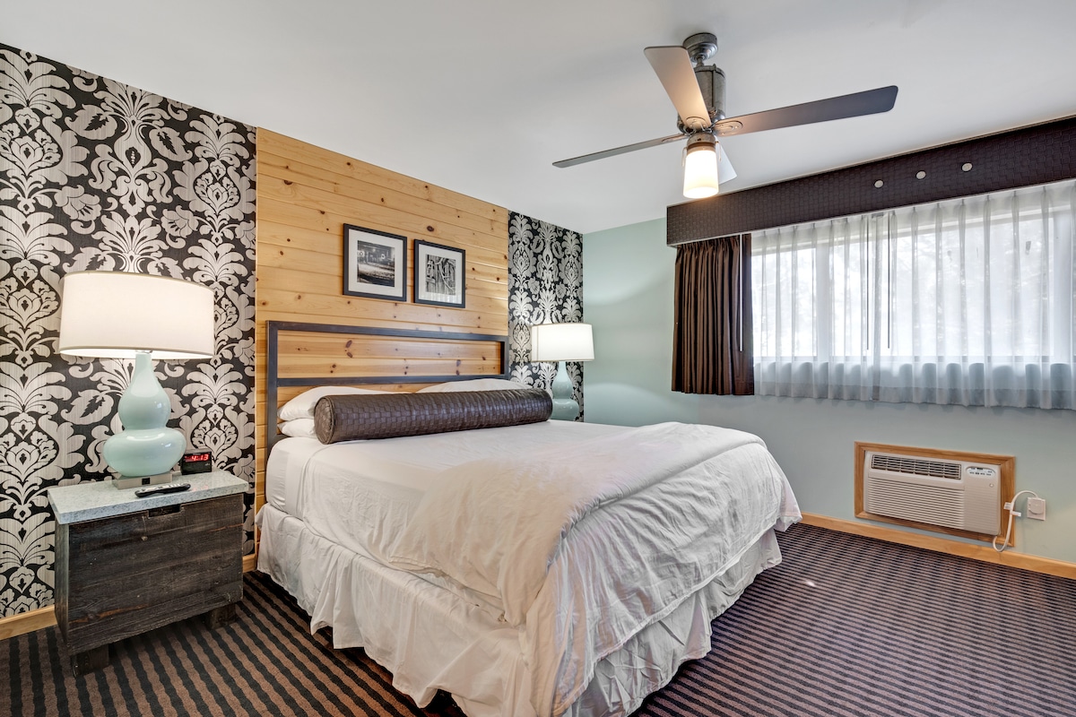 A well-appointed bedroom features a spacious bed layered with white bedding. Distinctive patterned wallpaper contrasts with wooden accents. A lighted ceiling fan and two bedside lamps provide illumination, while a large window dressed in sheer and dark fabric offers natural light.