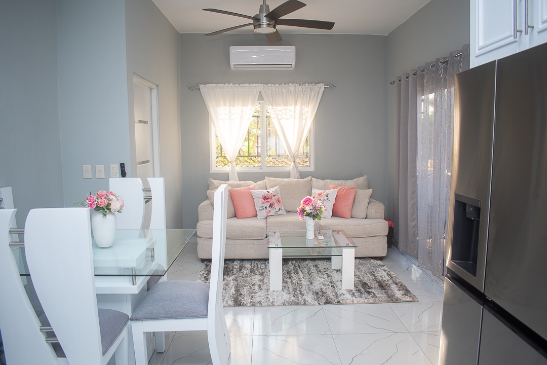 A bright living area features a comfortable sofa adorned with decorative pillows. A modern dining table is set with floral centerpieces. Light filters through sheer curtains, illuminating the stylish decor and polished marble flooring.