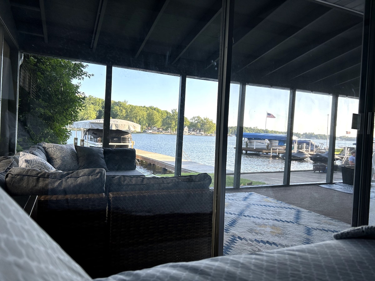 A serene view of Paw Paw Lake is captured from the screened-in porch, showcasing a spacious dock and boats moored nearby. Comfortable seating is arranged in the foreground, extending a welcoming atmosphere to enjoy the natural surroundings.