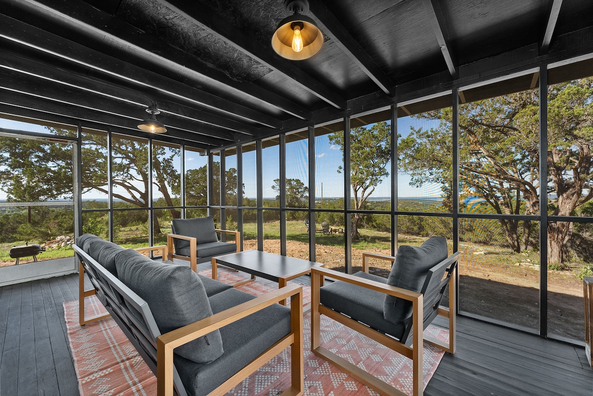A screened porch is presented with modern furnishings, featuring a wooden table surrounded by four comfortable armchairs. Large windows allow a view of the expansive outdoor landscape, where trees and rolling hills complement the serene setting.