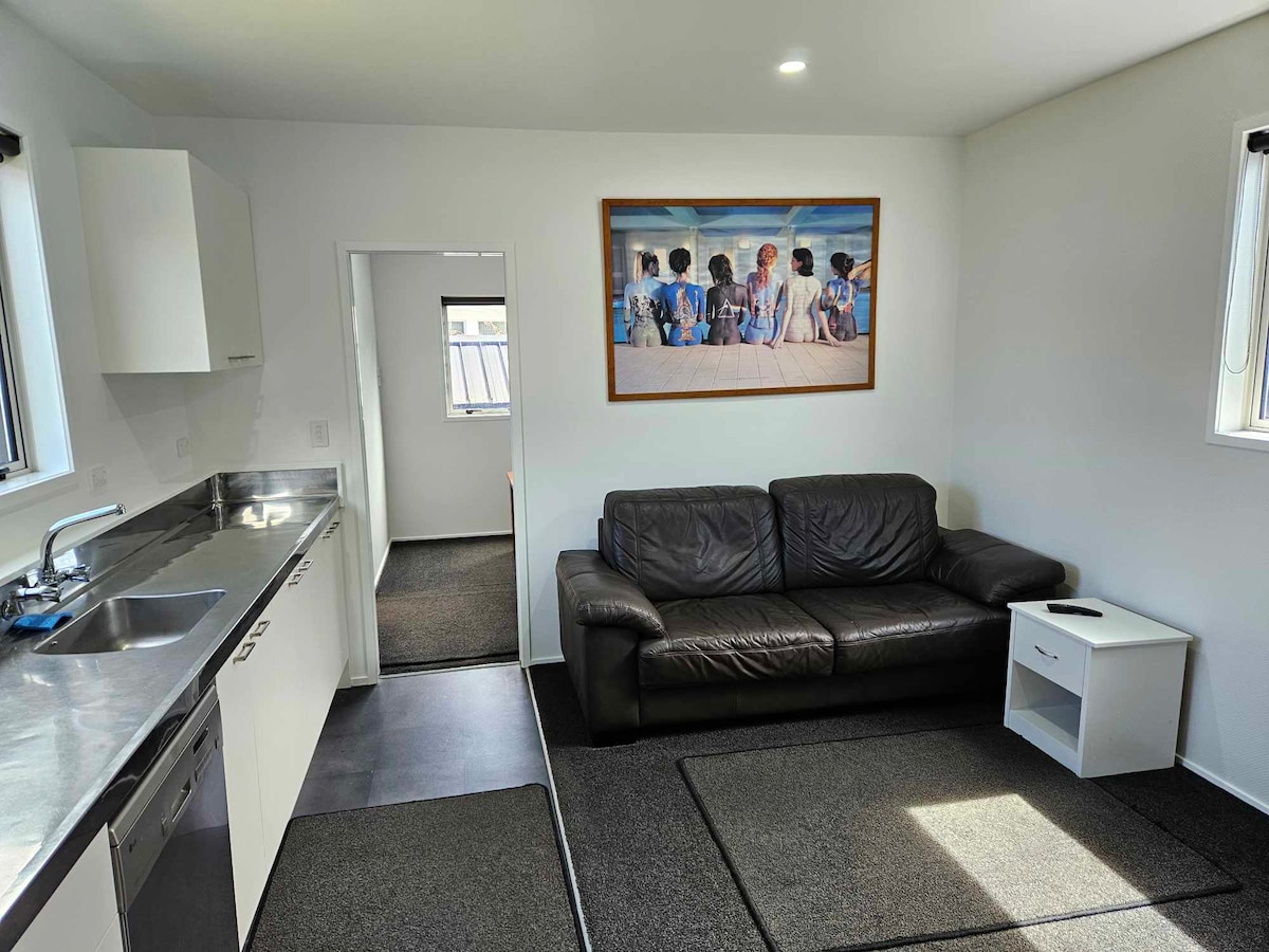 A modern lounge area features a dark leather couch paired with a small white side table. Light floods in through windows, illuminating the room. A large artwork depicting people is hung on the wall, adding visual interest to the space.