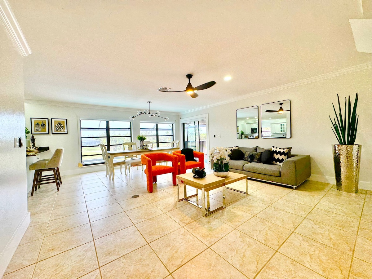 An open-concept living and dining area is showcased, featuring a comfortable gray sofa and vibrant red accent table. A dining table for six is positioned nearby, complemented by stylish decor and ample natural light from large windows.