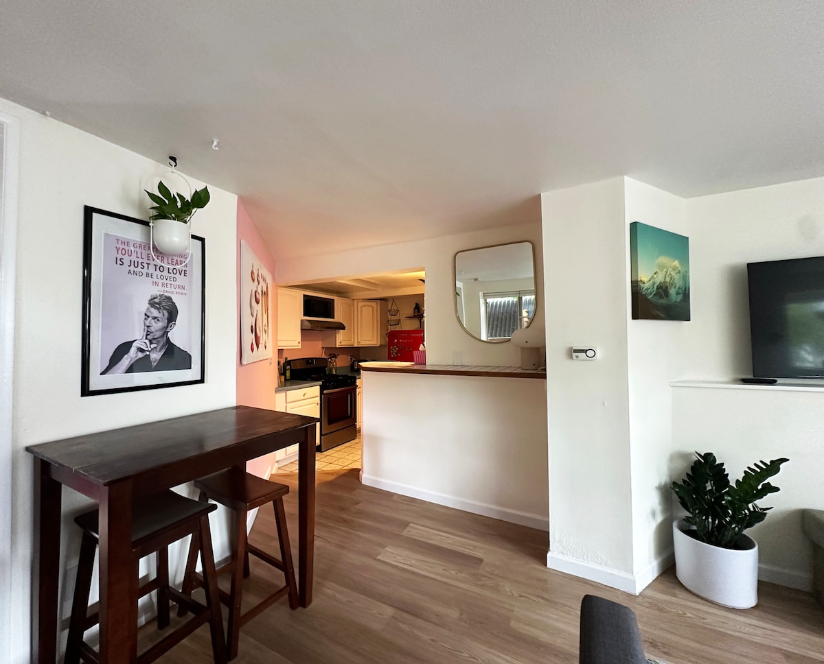 A bright living space features a small dining area with a table and two stools. In the background, a kitchen is visible, along with a mirror reflecting light. Artwork adorns the walls, while a potted plant is positioned in a corner, adding a touch of greenery.
