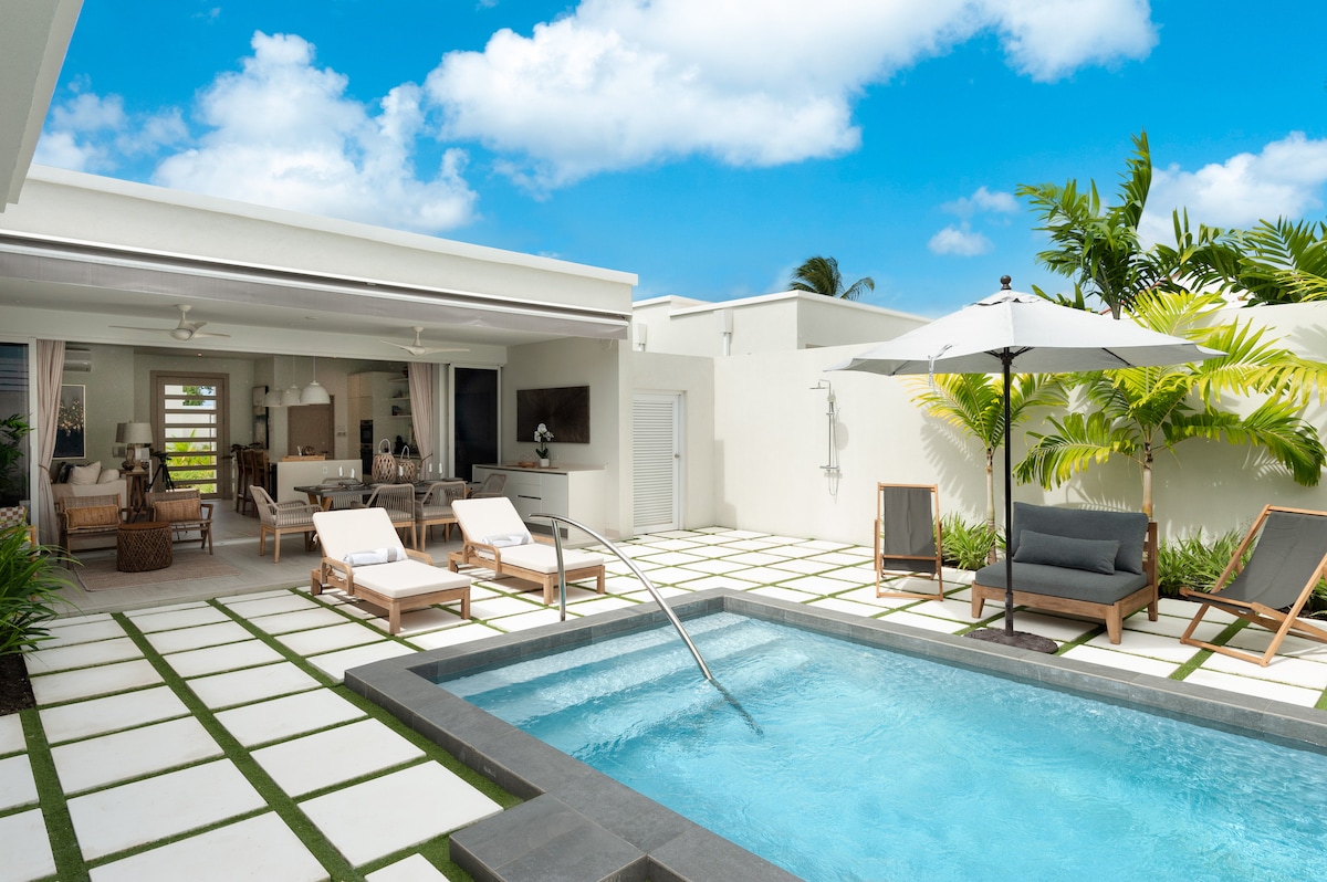 An inviting outdoor living space features a private pool surrounded by lounge chairs. A shaded seating area is placed under an umbrella, alongside tropical greenery. The spacious patio is paved with white tiles and accented by green grass patches, all beneath a clear blue sky.