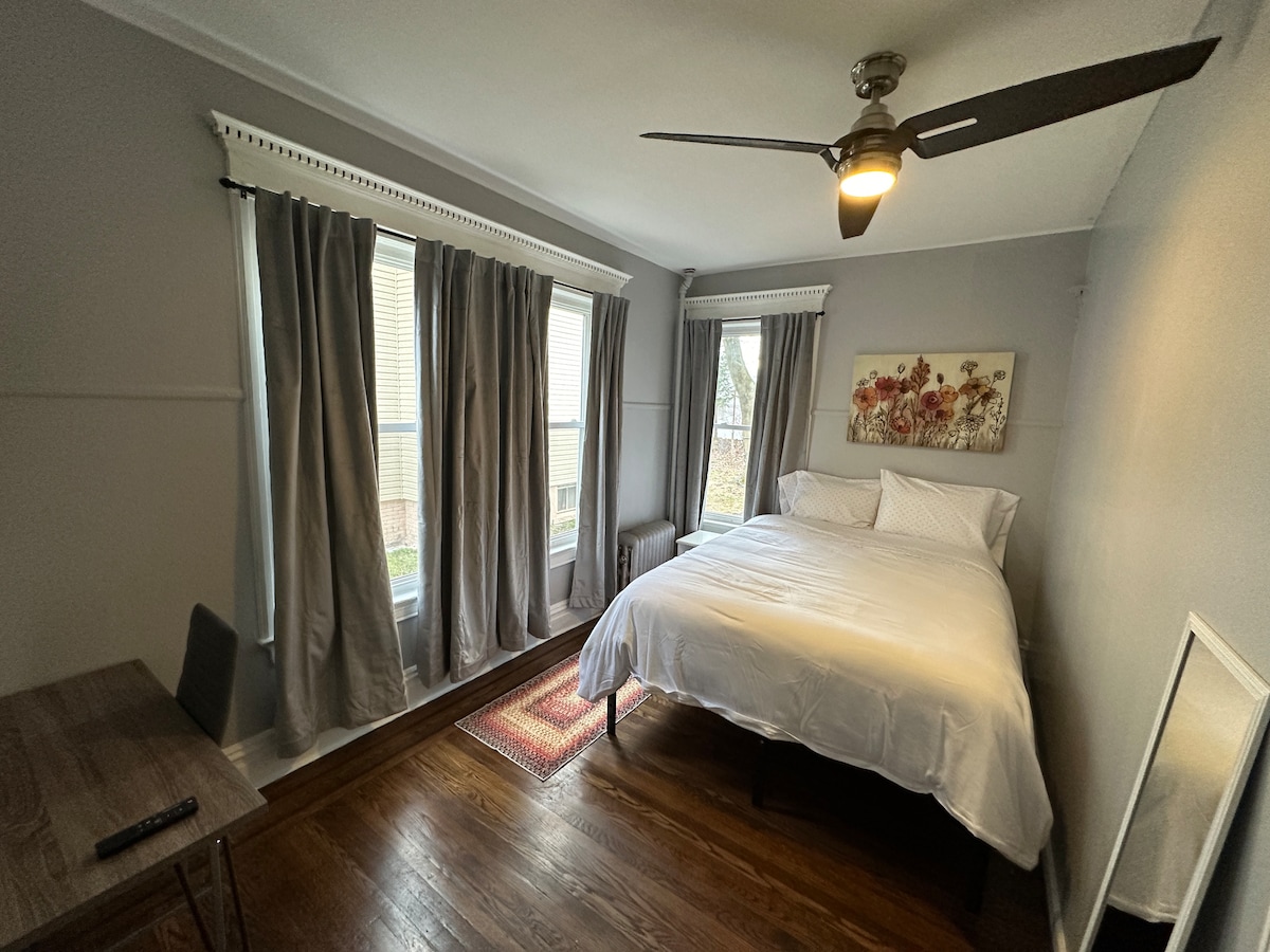A cozy bedroom features a spacious bed dressed in white linens, flanked by large windows draped with gray curtains allowing natural light to filter in. A small table and chair are positioned nearby, providing a dedicated workspace, while warm wooden flooring adds to the room's ambiance.