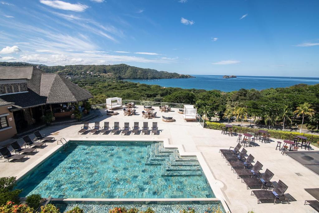 A spacious outdoor area is viewed from above, featuring a large pool with shimmering water and sun loungers arranged around it. Lush greenery surrounds the space, and a distant coastline is visible under a clear blue sky.
