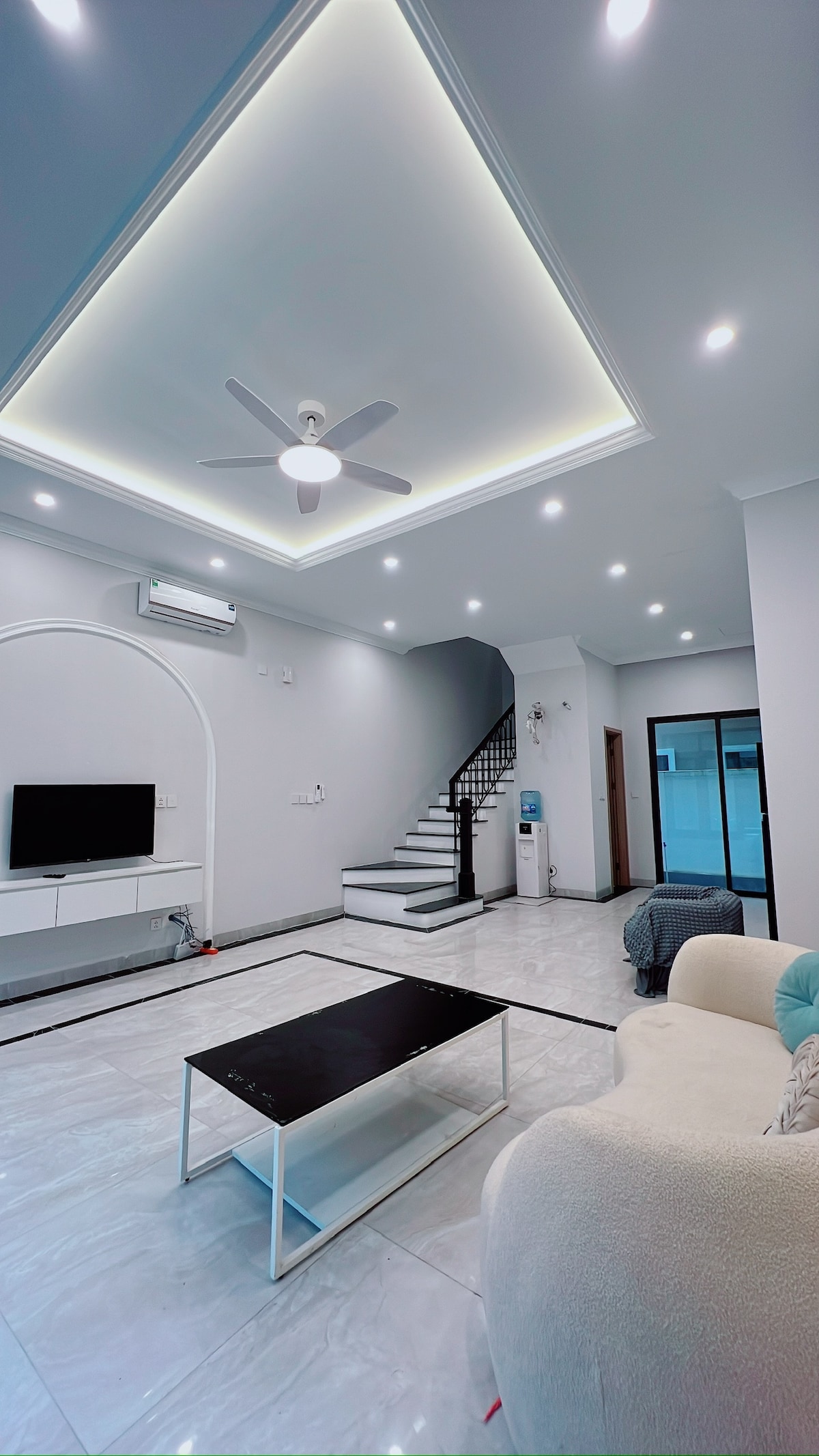 A modern living area is illuminated by recessed lighting, featuring a comfortable light-colored couch and a sleek black coffee table. A TV stands on a white cabinet, while a staircase with black and white steps leads to the upper level. Large windows allow natural light to fill the space.