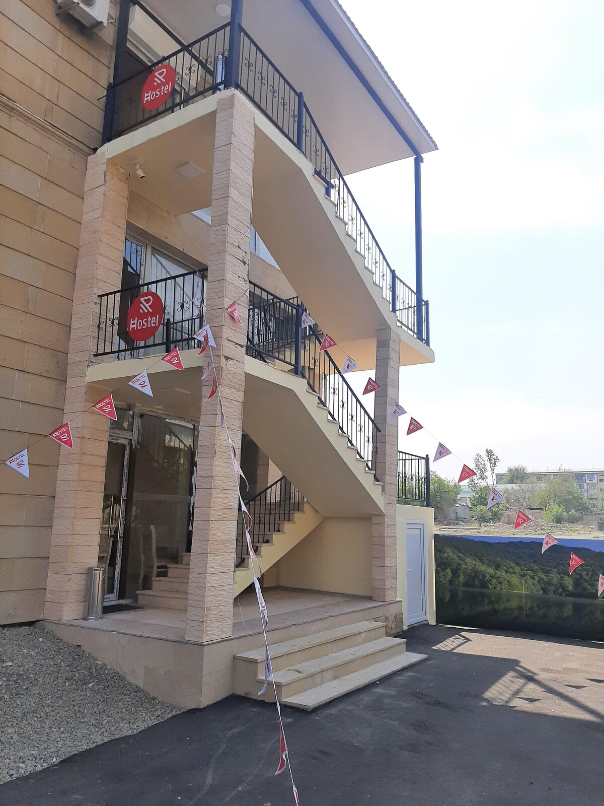 A three-story building is visible, featuring a staircase adorned with a black railing. Colorful flags are displayed along the railing, enhancing the entrance. Large glass doors lead to the main entry, and a partially visible outdoor area can be seen in the background.