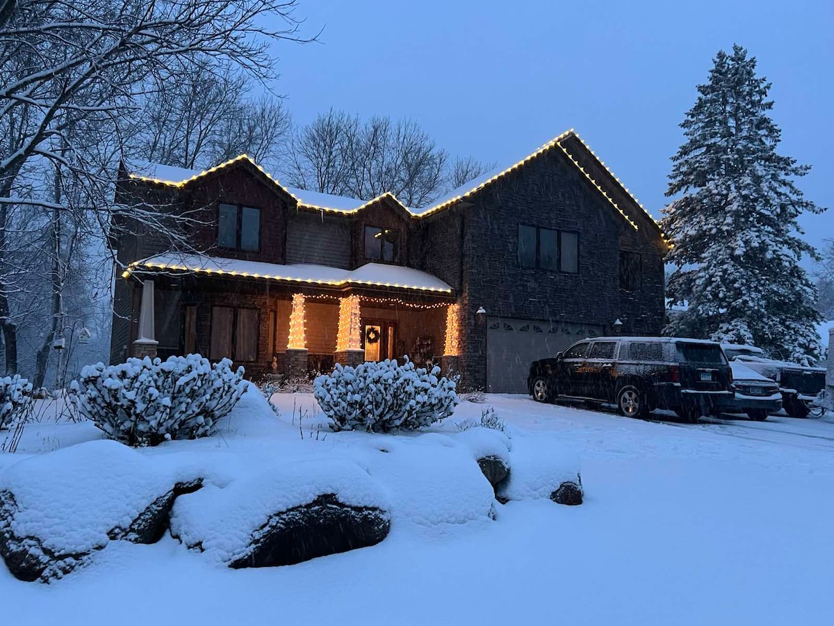 A structured home is outlined by warm lights against a snowy backdrop. Snow blankets the ground, with bushes and rocks visible in the front yard. Trees stand tall in the background, creating a serene winter atmosphere.