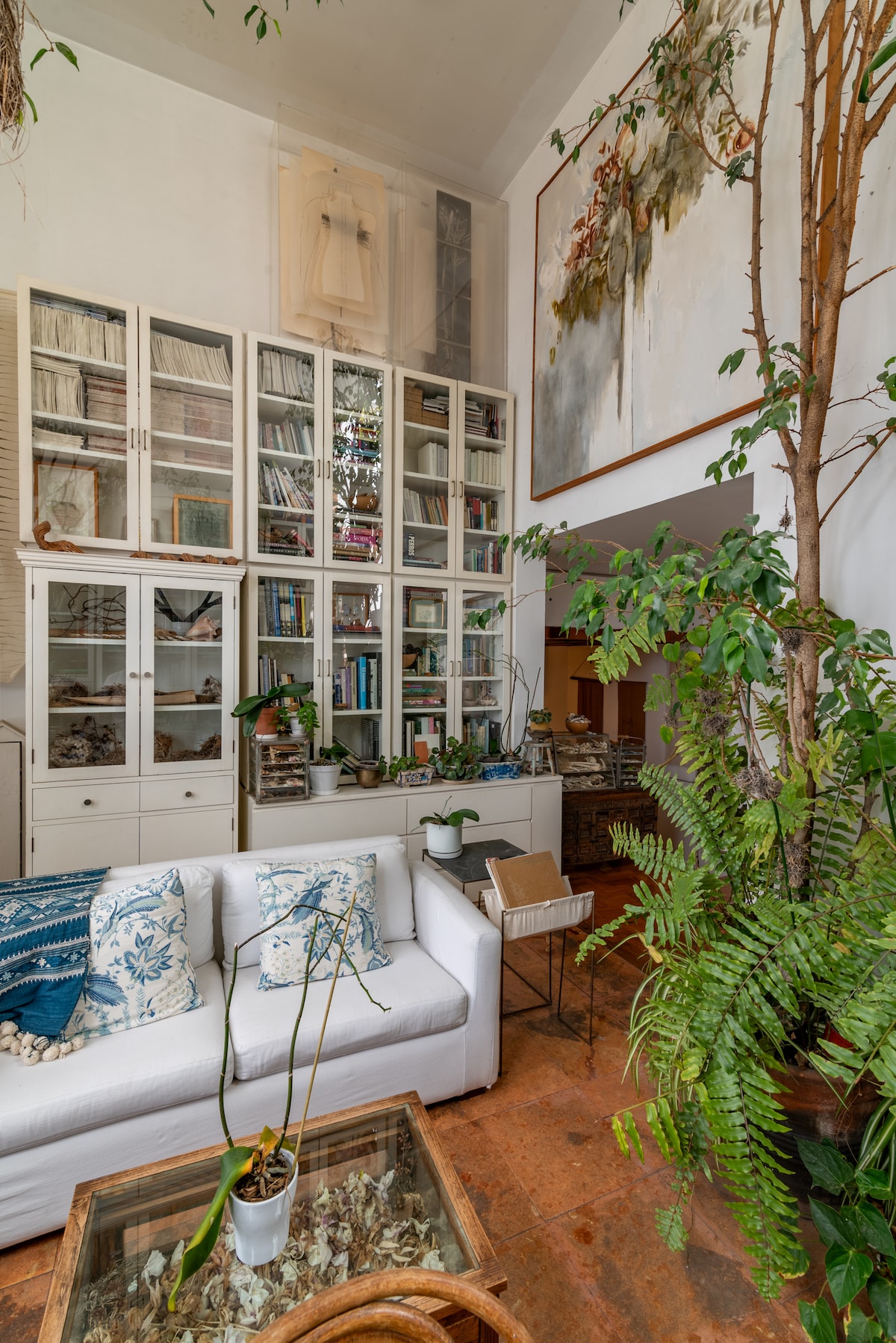 A light-filled living area features a white sofa adorned with decorative pillows, complemented by a glass coffee table. Tall shelves are lined with books and curated objects. A large piece of contemporary art hangs on the wall, surrounded by lush green plants, adding vibrancy to the space.