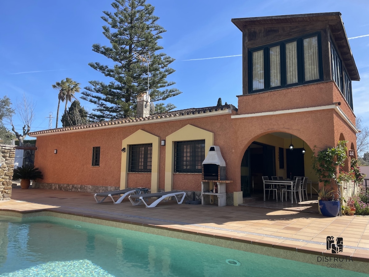 A charming holiday home features a swimming pool with a patio area. An outdoor dining space is available with a built-in barbecue nearby. Sun loungers are positioned around the pool. The building is adorned with large windows and a distinctive upper balcony, surrounded by landscaped greenery.
