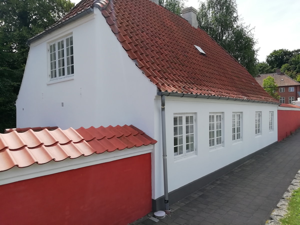 A charming white house is displayed with a red-tiled roof and matching red wall. Several windows, framed in white, allow ample natural light. The pathway leads alongside the house, providing access to the inviting entrance.