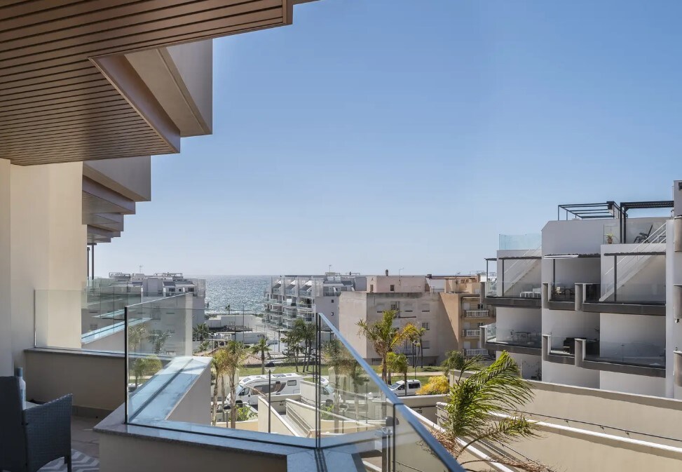 A spacious terrace features glass railings and views of the ocean in the distance. The landscape showcases modern buildings and palm trees, creating a relaxed atmosphere under a clear blue sky.