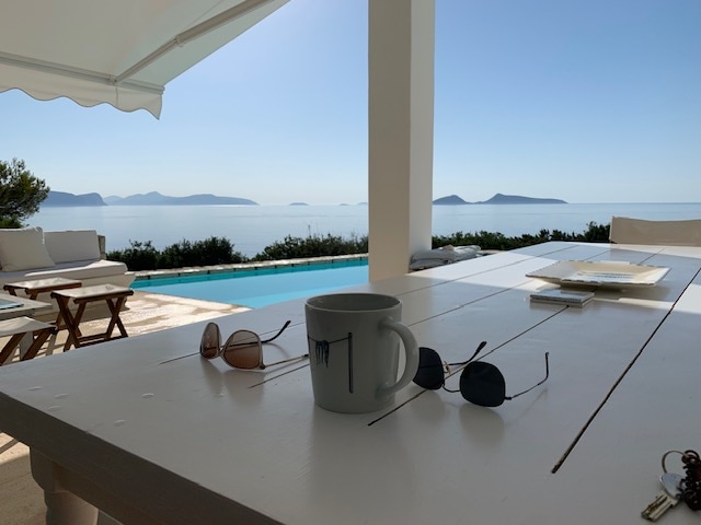 A table is set on a covered terrace, featuring a white surface with a cup and two pairs of sunglasses. In the background, a private pool reflects the bright sky, and distant islands are visible across the calm sea.