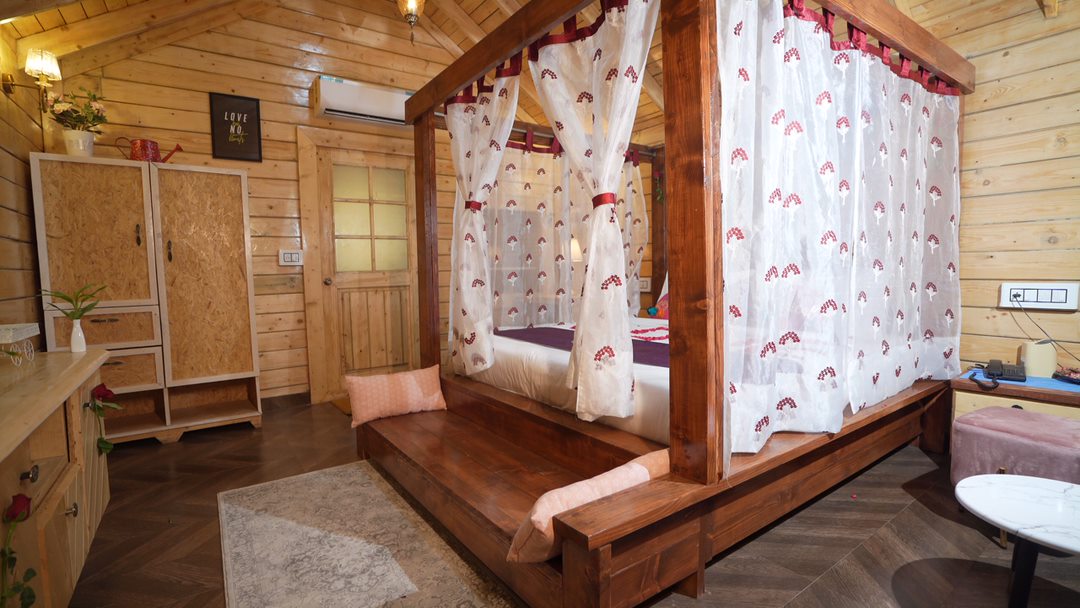 A wooden canopy bed with sheer curtains creates a cozy focal point in the room. Light-colored wooden walls add warmth, while a spacious wardrobe and a small seating area are visible. A soft area rug lies beneath, enhancing the inviting atmosphere.