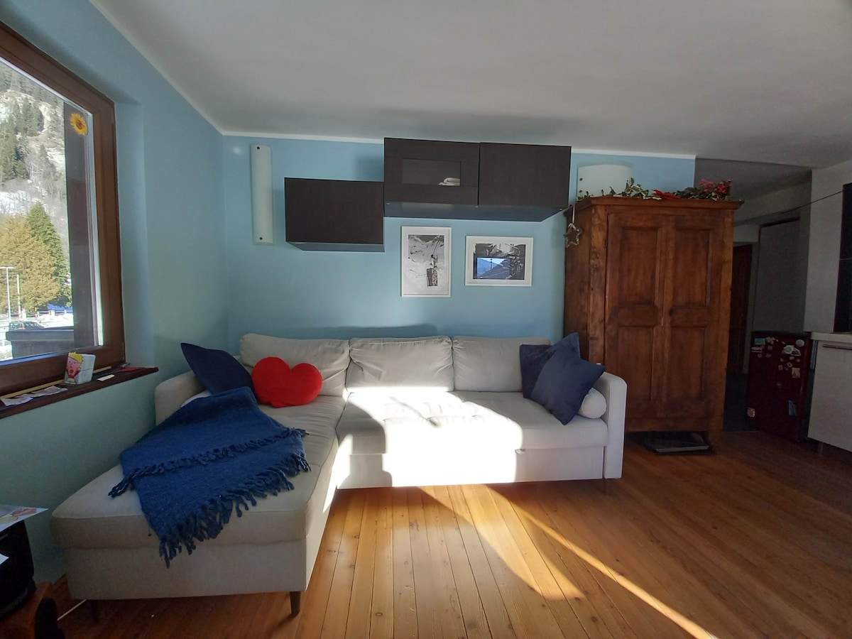 A light-filled living area features a spacious white sectional couch adorned with blue and navy cushions. A vibrant red heart pillow adds a pop of color. The wall displays framed images and has a wooden cabinet providing classic charm. Warm wood flooring complements the cozy atmosphere.