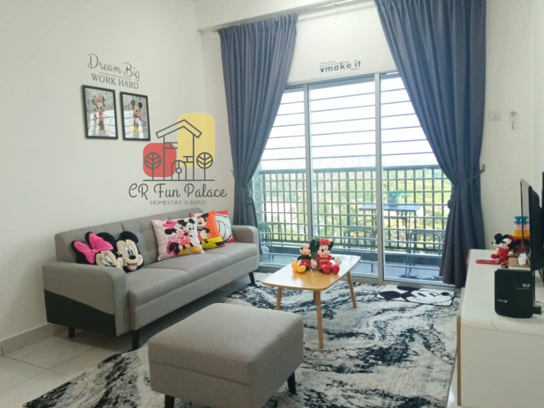 A bright living area features a comfortable sofa adorned with vibrant cushions, accompanied by a matching ottoman. Large windows dressed in dark curtains allow natural light to enter, while a patterned area rug complements the space. A television is positioned on a side table.