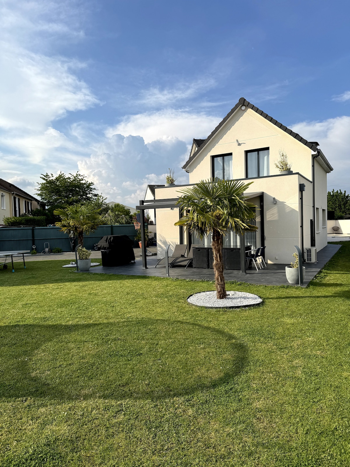 A modern house is set against a clear sky, featuring a well-maintained lawn with a palm tree and garden stones. A spacious patio area includes outdoor seating and is bordered by low fencing, offering a comfortable space for relaxation.