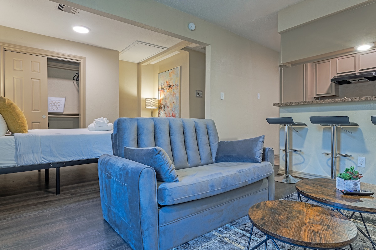 A furnished living area is showcased with a blue sofa and two circular tables. In the background, a spacious bed is partially visible, accompanied by a colorful wall art piece. The kitchen area reveals modern bar seating, enhancing the open-concept layout.