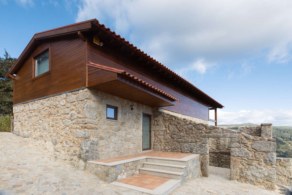 The exterior of the house is presented, showcasing a combination of stone and wooden materials. A sloped roof extends over the entrance, which is framed by stone steps. The surrounding landscape is visible, enhancing the natural setting.
