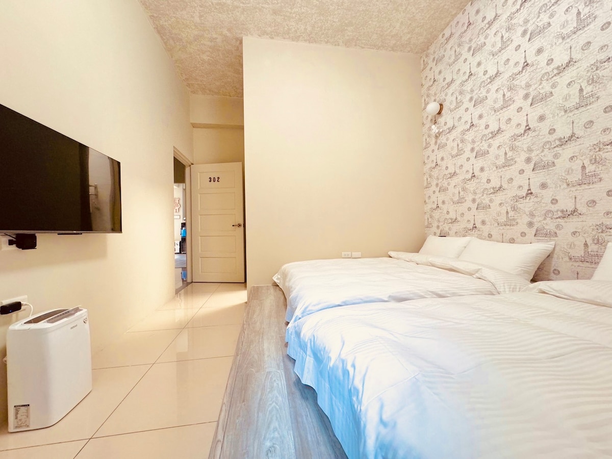 A spacious room features two neatly made beds with crisp white linens. A flat-screen television is mounted on the wall opposite the beds. Soft, neutral-colored walls complement the textured ceiling, and light is welcomed through the door leading to the hallway.