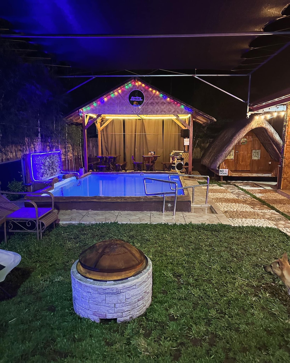 A relaxing outdoor area features a small pool surrounded by a grassy lawn. Colorful lights adorn a nearby hut, where seating is available. A fire pit with a stone surround adds a cozy touch to the space, which is framed by natural greenery.