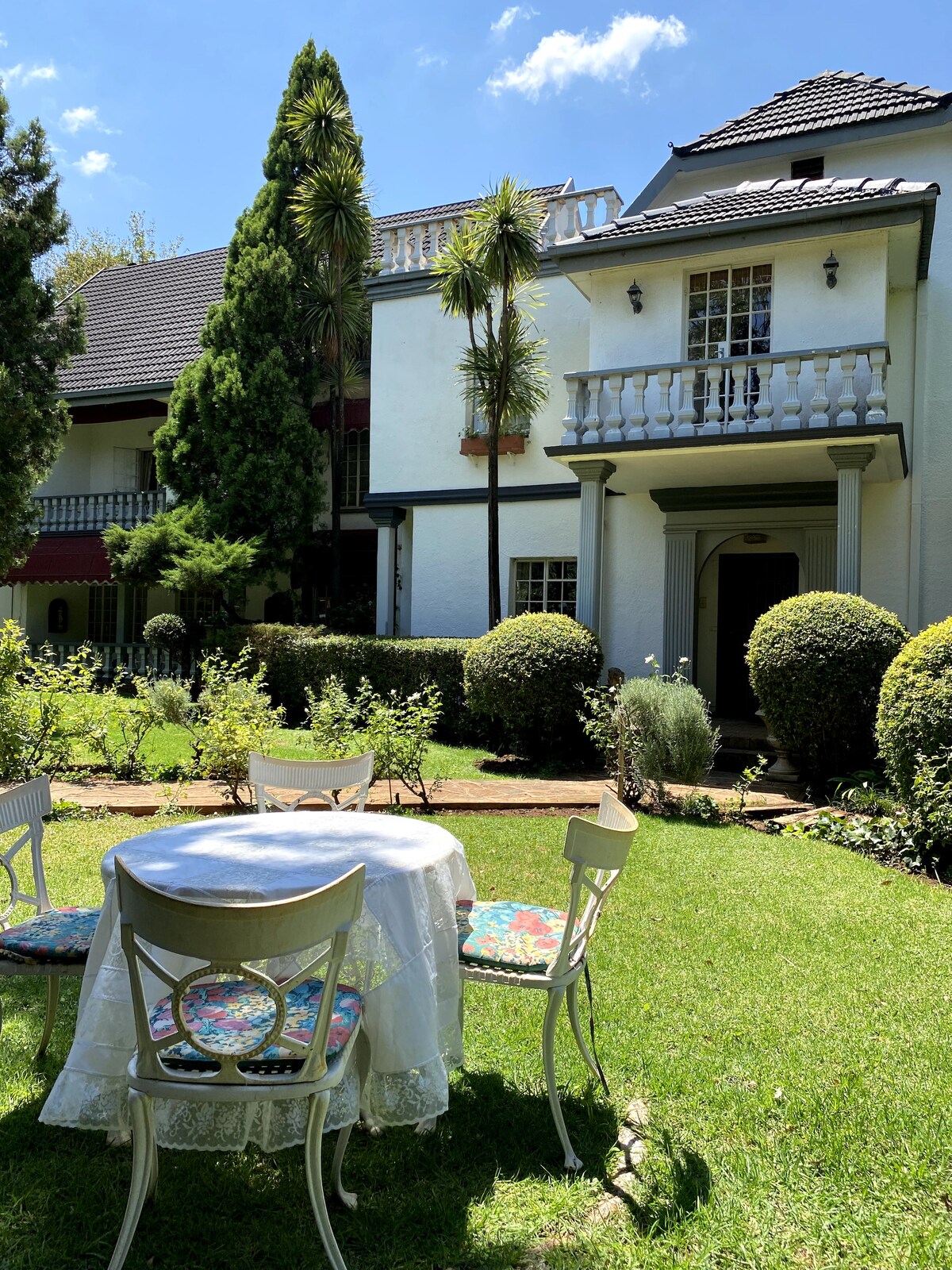 A charming outdoor area is highlighted by a circular table surrounded by four elegant chairs. Lush greenery, including neatly trimmed hedges and tall trees, frames the scene, while a two-story building with a balcony stands in the background, showcasing a blend of architectural styles.